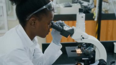 Laboratuvarda çalışırken cam kaydıraktaki numuneye mikroskopla bakan Afro-Amerikalı genç kadın biyolog.
