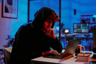 Siyah kapüşonlu, kıvırcık saçlı bir adam dizüstü bilgisayarını programlarken düşüncelerine dalmış.