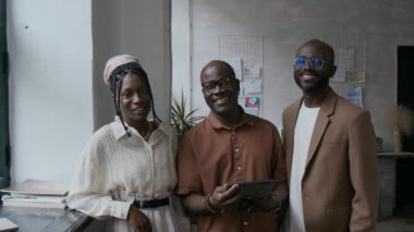 Üç iş arkadaşının portresi şık gündelik giysiler içinde birlikte duruyorlar, gülümsüyorlar ve modern ofiste kameraya poz veriyorlar.