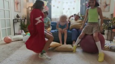 Kırmızı pelerinli çocuk ve kafasında karnaval bandı olan kız hopper topları üzerinde zıplarken üç arkadaş onları arka planda kanepede otururken izliyor.