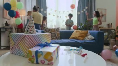 Hediye kutularını, parti şapkalarını ve ıslıklarını beyaz yuvarlak masaya yakın çekim, arka plana balon atan beş çocuk.
