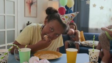 Parti şapkalı alfa kız arkadaşlar arka planda oynarken şenlik masasında pastanın tadını çıkarıyor.