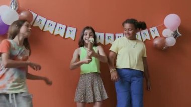 Doğum günü kızı patlıyor ve konfeti yağmurunun keyfini çıkarıyor. Arkadaşlarıyla şeftali rengi duvarlara karşı. Balonlarla süslenmiş.