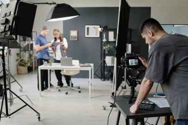 Yaratıcı yönetmeni ve asistanı sahne malzemelerini tartışırken tanınmayan genç kameraman kamera ayarlarını kontrol ediyor.