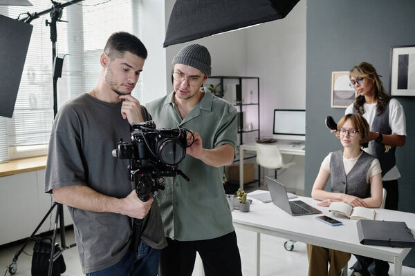 Молодой взрослый оператор и режиссер смотрит на дисплей камеры, обсуждая кадры, пока гримёры чистят зубы актрисе