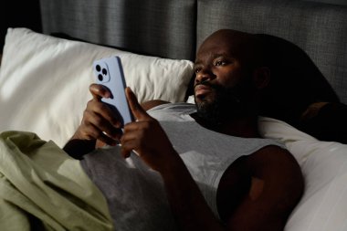 Afrika kökenli Amerikalı bir adamın yatakta uykusuzluk çekerken sosyal ağları kaydırdığı yüksek açılı bir çekim.