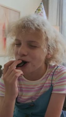 Pozitif gen alfa kızın dikey portresi. Sarı saçlı, çikolatalı kurabiye yiyen ve doğum günü kutlamasında kameraya gülümseyen.