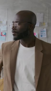 Kafasını çevirip kameraya gülümseyen ve ofisinde dikilen Afro-Amerikan bir adamın dikey portresi.