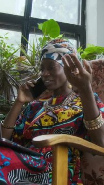 Siyahi kadının geleneksel Afrika kıyafeti ve aksesuarlarıyla telefonda konuşurken, sürdürülebilir bir ofiste koltukta rahat bir şekilde otururken, dikey düşük açılı görüntüsü.