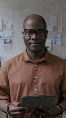Afro-Amerikan üst düzey bir adamın dijital tableti tutarken ve modern ofiste dururken gülümseyerek kameraya bakarken çekilmiş dikey portresi.