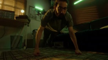 Sakallı adam, loş ışıklı, neon yeşil ışıklı retro odasında yerde parmak şınavı çekiyor.