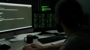 Sakallı programcı yoğun bir şekilde çift monitörlere odaklanıyor loş ışıklı odada yeşil ve beyaz kod çizgileri gösteriyor, geceleri çalışıyor.