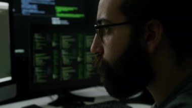 Sakallı programcının bilgisayar ekranının önünde oturmuş, odaklanmış ifadesiyle yoğun bir şekilde kodun birden fazla monitörde çalışan satırlarını izlerken yakın plan görüntüsü.
