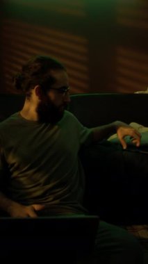 Sakallı bir adamın dizüstü bilgisayarıyla çalışırken ve akıllı telefondan mesaj atarken loş bir odada otururken dikey görüntüsü.