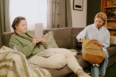 Down sendromlu bir kız kanepeye oturmuş, annesi okul için sırt çantası hazırlarken dijital tablet ekranlara bakıyor.
