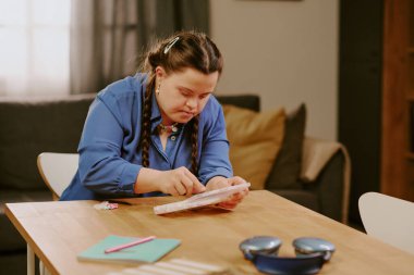 Caucasian girl with Down syndrome spending spare time at home, sitting at table and beading bracelet clipart