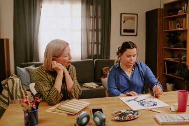 Oturma odasında oturan olgun bir kadın Down sendromlu kızının resim çizmesini izliyor.