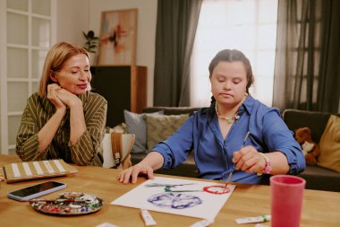 Mature Caucasian woman sitting at desk in living room, enjoying watching her daughter with disability painting flower on paper clipart