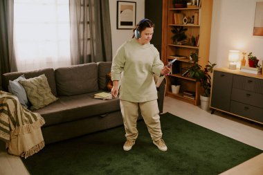 Down sendromlu genç bir kadının günlük giysiler ve kulaklıklarla oturma odasında durup akıllı telefondan müzik listesi seçerken geniş açılı görüntüsü.