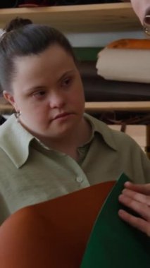 Down sendromlu bir gencin dikey çekimi. Deri atölyesinde usta bir zanaatkâr tarafından tutulan kumaşı inceliyor.