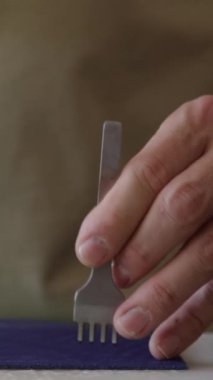 Tanımlanamayan ellerin dikey yakın çekimi keskinin çatallı ucunu stüdyodaki tezgahta çekiçle koyu mavi deri parçasına bastırarak dikişler oluşturur.