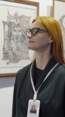 Kırmızı saçlı, gözlüklü, rozetli, sanat galerisinde yürüyen ve sanat eserlerini inceleyen bir kadının dikey görüntüsü.
