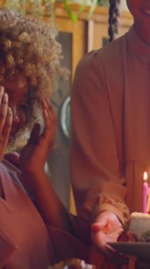 Dikey mutlu zenci kadın pastaya mum üflerken ve evde ailesiyle doğum gününü kutlarken genç kıza sarılırken.