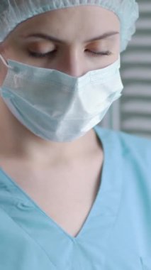 Tıbbi maske takan, önlük takan ve klinikte kameraya bakan hemşirenin dikey portresi.