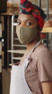 Önlük, başörtüsü ve koruyucu yüz maskesi takan, fırında ekmek vitrininin yanında duran ve kameraya poz veren genç siyahi kadının dikey portresi.