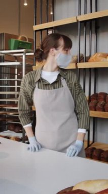 Önlük, maske ve eldiven takan kadın fırın işçisinin müşteriden sipariş alırken, sonra da kağıt torbaya taze ekmek koyup satarken dikey pozu.