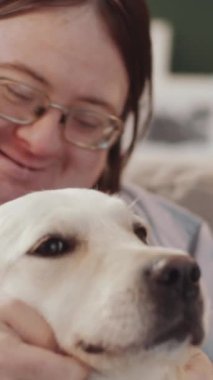 Down sendromlu gülümseyen bir adamın dikey yakın çekimi. Evde oturmuş, tatlı Golden Retriever köpeğini nazikçe okşuyor.