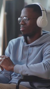 Tekerlekli sandalyede, kablosuz kulaklıkla müzik dinleyen ve kameraya gülümseyerek poz veren genç Afrikalı Amerikalı bir adamın dikey portresi.