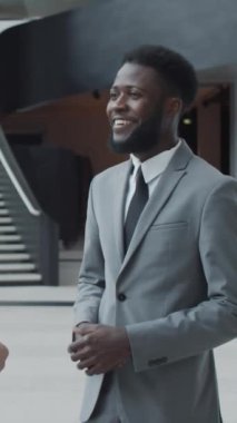 Resmi takım elbiseli genç siyahi işadamının modern ofis binasındaki meslektaşıyla gülümseyip konuştuğu dikey bir fotoğraf.