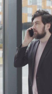 Kafkasyalı sakallı, şık giyinmiş, ofis binasında koridorda yürüyen ve cep telefonuyla konuşan beyaz iş adamının dikey görüntüsü.