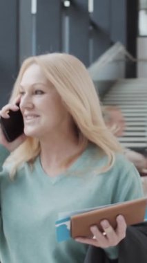 Orta yaşlı sarışın bir kadının elinde pasaportuyla havaalanında yürürken çekilmiş dikey bir fotoğrafı.