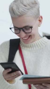 Dikey çekimde, kısa saçlı genç bir kadın havaalanında duruyor, elinde uçuş kartı, cep telefonuyla mesaj atıyor ve gülümsüyor, sonra da kapıyı ararken etrafa bakıyor.