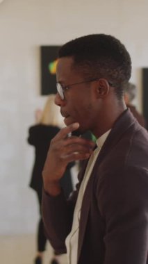 Gözlüklü Afrikalı Amerikalı bir gencin dikey görüntüsü. Tabloyu dikkatle inceliyor. Sanat galerisinde çenesini ovuyor.