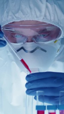 Koruyucu giysiler giymiş genç erkek bilim adamının dikey yakın görüntüsü kırmızı maddeli pipeti test tüpüne koyup ilaç araştırması yaparken içini dolduruyor.
