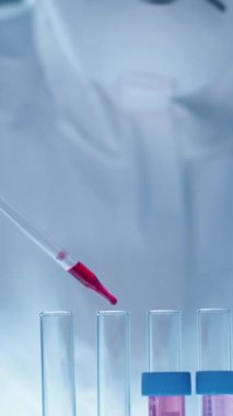 Koruyucu üniformalı genç bir kadın kimyagerin zehirli çözücüleri analiz ederken kırmızı çözeltiyi test tüpüne dökmesini dikey olarak yukarı doğru eğ.