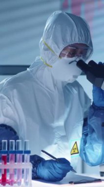 Çağdaş laboratuarda mikrobiyoloji araştırması yaparken mikroskopla çalışan ve sonuçları yazan kadın bilim adamının dikey görüntüsü