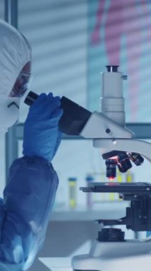 Modern laboratuarda çalışırken koruyucu ekipman giyip mikroskop altında örnekleri inceleyen kadın epidemiyolojistin dikey görüntüsü
