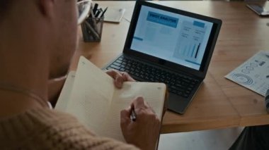 Defterine notlar alan bir adamın omuz üstü fotoğrafı modern ofisteki iş toplantısında sunumu dikkatle dinlerken.