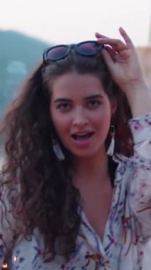 Kafkasyalı esmer kızın dikey portresi. Fotoğraf makinesi için dans ediyor ve arkadaşlarıyla açık hava partisinde güneş gözlüğü takıyor.