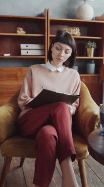 Genç esmer kadın danışmanın portresi dikey zum. Panoya gelişigüzel yazılar yazıp modern ofiste koltukta otururken kameraya gülümsüyor.