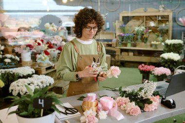 Kadife önlük giymiş kıvırcık saçlı kadın iş masasının yanında dizüstü bilgisayarlı ve farklı tipte çiçeklerle buket yapıyor.