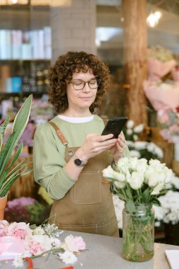 Genç beyaz çiçekçinin elinde akıllı telefon tutarken ve sosyal ağları kontrol ederken çiçekçide mola verirken dikey çekimleri.
