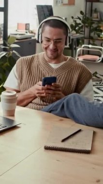 Kulaklıkla müzik dinleyen, akıllı telefonuyla sosyal medyada kayan, modern ofiste kahve molası veren genç bir adamın dikey görüntüsü.