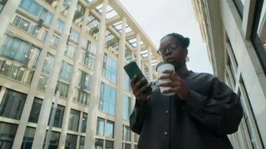 Güneşli bir günde modern şehirde yürürken akıllı telefonuna bakıp kahve içen Afrikalı Amerikalı kadının düşük açılı çekimi.