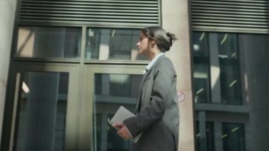 Resmi takım elbiseli genç melez kadın CEO 'nun elinde akıllı telefon ve defterle modern cam cepheli binanın önünden geçerken düşük açılı izleme görüntüsü.
