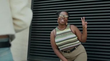 Yazlık kıyafetli siyahi kadın geniş bir gülümseme içinde, siyah duvarın önünde poz verirken barış işareti yapıyor.